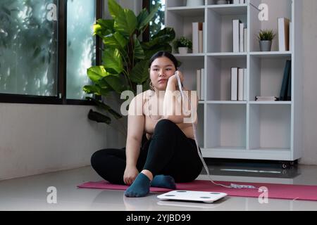 Jeune femme assise sur un tapis de yoga avec ruban à mesurer et échelle dans un intérieur moderne avec des plantes vertes et une étagère à livres Banque D'Images