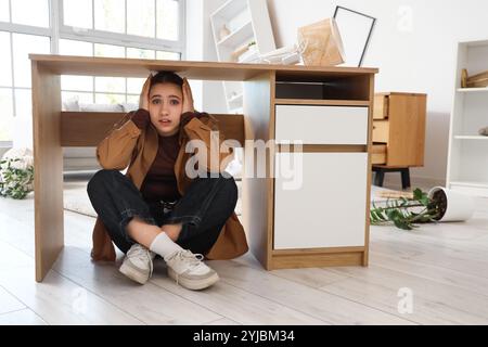 Jeune femme effrayée se cachant sous la table à la maison pendant le tremblement de terre Banque D'Images
