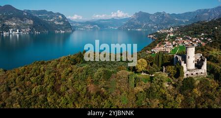 Vue aérienne d'une tour de pierre historique sur une colline luxuriante surplombant un lac bleu serein, entouré de montagnes et d'un charmant village au bord du lac. Banque D'Images