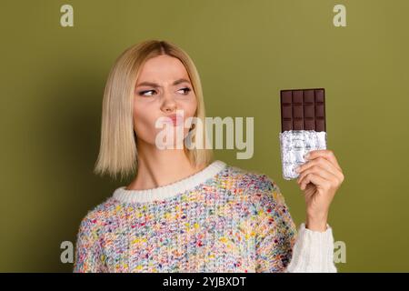 Jeune femme montrant une barre de chocolat avec une expression humoristique devant un fond kaki favorisant l'indulgence douce Banque D'Images