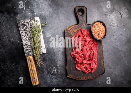 Viande de boeuf crue non cuite tranchée, lanières pour la cuisson du bœuf stroganoff sur une planche de bois. fond noir. vue de dessus Banque D'Images
