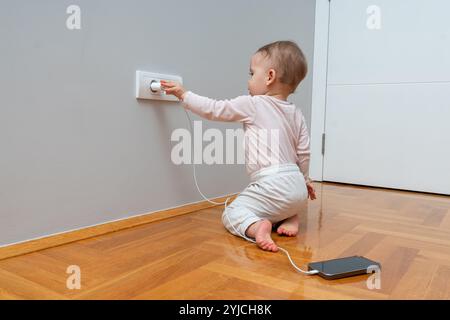 Un tout-petit curieux explorant une prise électrique avec chargeur de téléphone, soulignant l’importance de la sécurité à la maison et de la vigilance parentale. Banque D'Images