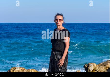 Debout sur des rivages rocheux, une femme regarde l'océan tranquille, trempant dans le soleil et la sérénité. Banque D'Images