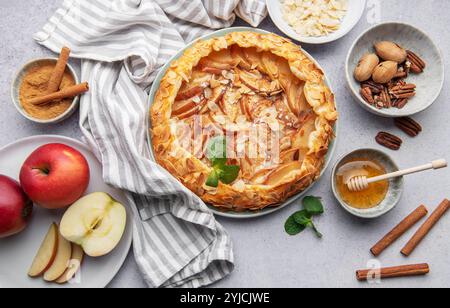 Une tarte aux pommes magnifiquement présentée ornée d'amandes et de miel se trouve sur une table rustique. Autour de lui se trouvent des pommes fraîches, des bâtonnets de cannelle et des bols Banque D'Images