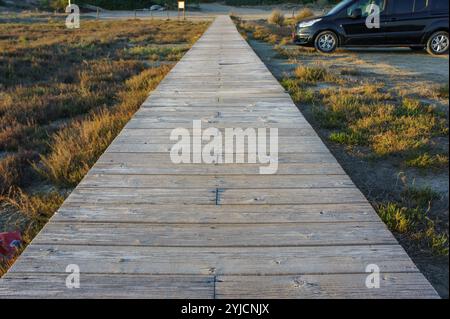 Un long chemin en bois mène à travers de hautes herbes, avec un véhicule garé à côté près de la mer tranquille. Banque D'Images