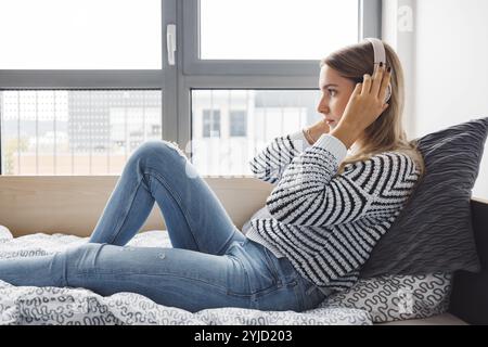 Blonde jeune femme caucasienne allongée sur son lit de dortoir à côté d'une fenêtre mettant des écouteurs, écoutant de la musique un jour d'automne froid et nuageux Banque D'Images