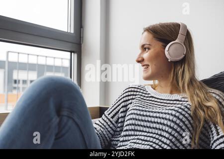 Blonde jeune femme caucasienne allongée sur son lit de dortoir à côté d'une fenêtre mettant des écouteurs, écoutant de la musique un jour d'automne froid et nuageux Banque D'Images