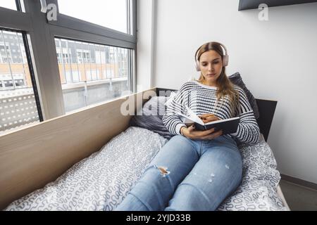 Blonde jeune femme caucasienne allongée sur son lit de dortoir à côté d'une fenêtre mettant des écouteurs, écoutant de la musique un jour d'automne froid et nuageux Banque D'Images