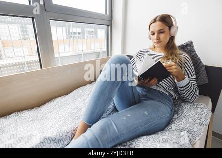 Blonde jeune femme caucasienne allongée sur son lit de dortoir à côté d'une fenêtre mettant des écouteurs, écoutant de la musique un jour d'automne froid et nuageux Banque D'Images