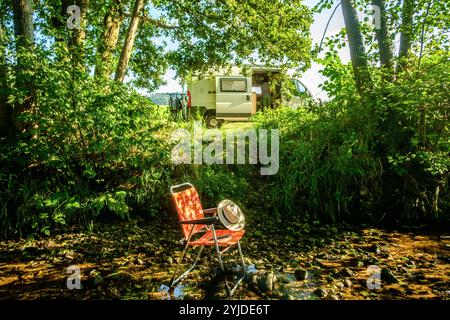 Ein Campingstuhl auf dem ein Buch und ein Stohhut liegt steht in einem Flussbett auf einem schattigen Platz unter einem Baum, im Hintergrund ist ein Wohnmobil zu sehen. Elsass, Frankreich. Ein Campingstuhl steht im Schatten in einem Fluss *** Une chaise de camping avec un livre et un chapeau de paille sur elle se dresse dans un lit de rivière dans un endroit ombragé sous un arbre, en arrière-plan un camping-car peut être vu Alsace, France Une chaise de camping se dresse à l'ombre dans une rivière Banque D'Images