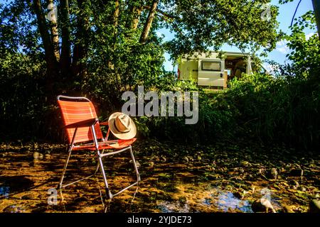 Ein Campingstuhl auf dem ein Buch und ein Stohhut liegt steht in einem Flussbett auf einem schattigen Platz unter einem Baum, im Hintergrund ist ein Wohnmobil zu sehen. Elsass, Frankreich. Ein Campingstuhl steht im Schatten in einem Fluss *** Une chaise de camping avec un livre et un chapeau de paille sur elle se dresse dans un lit de rivière dans un endroit ombragé sous un arbre, en arrière-plan un camping-car peut être vu Alsace, France Une chaise de camping se dresse à l'ombre dans une rivière Banque D'Images