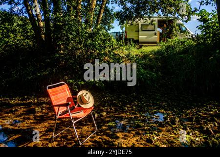 Ein Campingstuhl auf dem ein Buch und ein Stohhut liegt steht in einem Flussbett auf einem schattigen Platz unter einem Baum, im Hintergrund ist ein Wohnmobil zu sehen. Elsass, Frankreich. Ein Campingstuhl steht im Schatten in einem Fluss *** Une chaise de camping avec un livre et un chapeau de paille sur elle se dresse dans un lit de rivière dans un endroit ombragé sous un arbre, en arrière-plan un camping-car peut être vu Alsace, France Une chaise de camping se dresse à l'ombre dans une rivière Banque D'Images