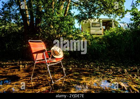 Ein Campingstuhl auf dem ein Buch und ein Stohhut liegt steht in einem Flussbett auf einem schattigen Platz unter einem Baum, im Hintergrund ist ein Wohnmobil zu sehen. Elsass, Frankreich. Ein Campingstuhl steht im Schatten in einem Fluss *** Une chaise de camping avec un livre et un chapeau de paille sur elle se dresse dans un lit de rivière dans un endroit ombragé sous un arbre, en arrière-plan un camping-car peut être vu Alsace, France Une chaise de camping se dresse à l'ombre dans une rivière Banque D'Images