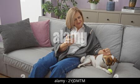 Une femme mature aux cheveux blonds se détend dans son salon avec son petit chien, en utilisant son téléphone sur un canapé confortable. Banque D'Images