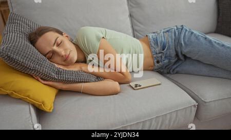 Une belle jeune femme dort paisiblement sur un canapé gris à la maison, avec un smartphone couché à côté d'elle, portant un Jean décontracté et un haut léger. Banque D'Images