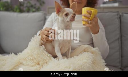 Une femme caucasienne mature est assise dans son salon confortable, tenant une tasse et caressant son petit chien chihuahua assis sur une couverture moelleuse. Banque D'Images