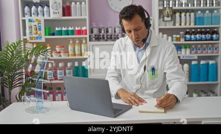 Homme hispanique d'âge moyen travaillant dans une pharmacie portant une blouse de laboratoire et un casque, écrivant sur un bloc-notes à un bureau blanc avec un ordinateur portable et des lunettes. Banque D'Images