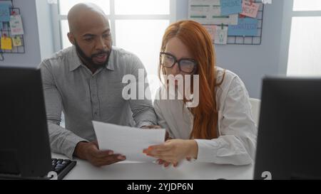 Homme et femme collaborant dans un bureau, examinant un document à un bureau avec des ordinateurs et un tableau de stratégie sur le mur. Banque D'Images