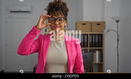 Jeune femme portant des lunettes et un blazer rose debout dans une salle de bureau ajustant ses lunettes avec une expression confiante Banque D'Images