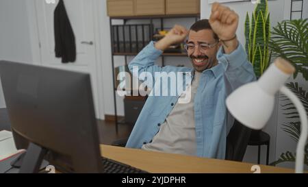 Jeune homme hispanique avec moustache célébrant dans un espace de travail de bureau avec une expression joyeuse. Banque D'Images