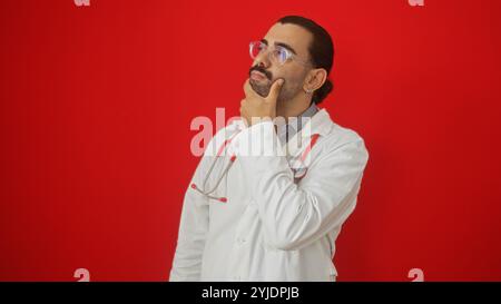 Jeune homme hispanique avec une moustache et des lunettes portant une blouse de laboratoire blanche et un stéthoscope rouge sur un fond rouge isolé, regardant réfléchi Banque D'Images