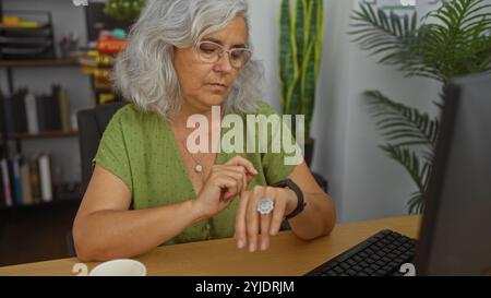 Femme portant des lunettes avec des cheveux gris dans une blouse verte regardant smartwatch dans le bureau avec des plantes et des étagères Banque D'Images