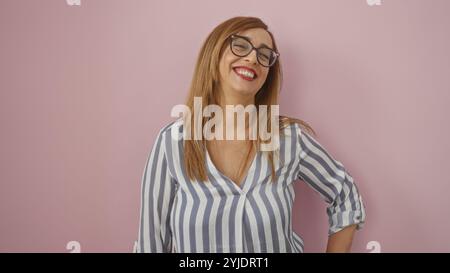 Femme souriant avec confiance, debout contre un mur rose, d'âge moyen avec des cheveux blonds et des lunettes, fond rose isolé, chemisier rayé, fe mature Banque D'Images