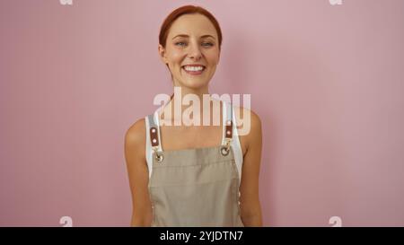 Jeune femme aux cheveux roux souriant devant un mur rose, portant un tablier beige avec des lanières de cuir sur ses épaules Banque D'Images