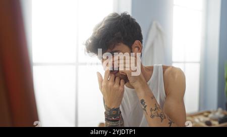 Un jeune homme hispanique avec une barbe et des tatouages, portant un débardeur blanc, serre un bouton sur son visage dans une salle de spa intérieure lumineuse. Banque D'Images