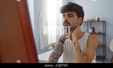 Beau jeune homme hispanique avec une barbe et des tatouages, assis dans une salle de spa tranquille, réfléchissant sur son parcours de bien-être, créant une atmosphère sereine et introsp Banque D'Images