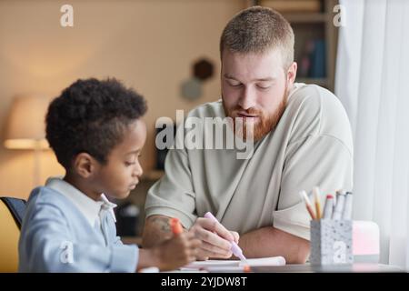 Photo moyenne d'un papa de soutien aidant son petit fils adopté à faire ses devoirs tout en étant assis au bureau et dessinant avec des stylos feutres colorés dans la chambre des enfants Banque D'Images