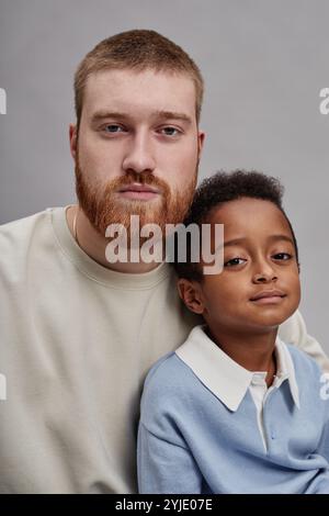 Portrait vertical de l'homme sérieux avec la barbe rouge posant à côté du petit garçon noir adopté pour la photo de famille en studio sur fond gris Banque D'Images