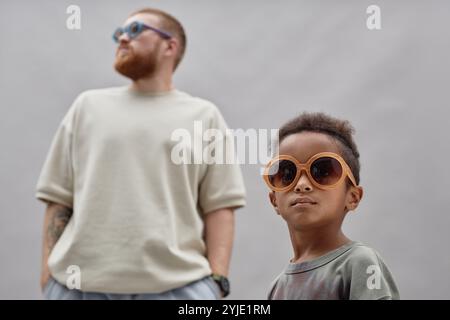 Plan moyen de petit garçon noir élégant dans de grandes lunettes de soleil regardant loin avec père adoptif debout en arrière-plan portant ressemblant à l'accessoire agai Banque D'Images