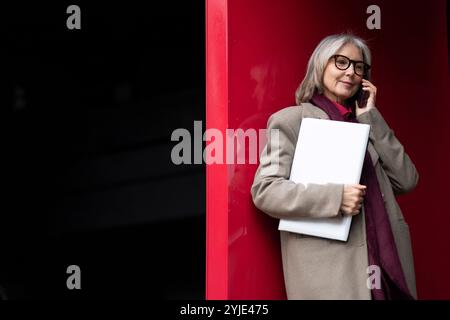 Femme d'affaires âgée dans un manteau élégant se tient en toute confiance, tenant un ordinateur portable tout en parlant sur son téléphone dans un cadre urbain moderne Banque D'Images