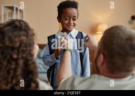 Plan moyen de petit garçon noir joyeux avec un sourire franc se préparant pour l'école pendant que les parents aident l'enfant à ajuster la chemise uniforme dans la chambre, copie Banque D'Images