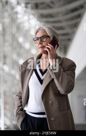 Une femme plus âgée dans un manteau élégant engagé dans un appel téléphonique tout en se tenant debout dans un bâtiment moderne à l'architecture frappante Banque D'Images