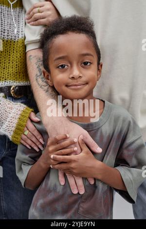 Portrait vertical d'un garçon noir adopté heureux tenant la main de papa posant pour la caméra avec des parents soutenant debout derrière l'enfant dans le studio Banque D'Images