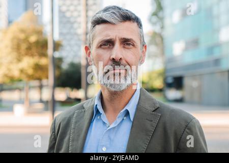 Homme d'affaires mature confiant regardant la caméra avec une expression sérieuse, homme professionnel avec la barbe dans une veste de costume debout à l'extérieur dans un cadre urbain, exsudant le leadership et l'expérience. Photo de haute qualité Banque D'Images