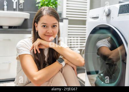 Jeune femme attendant que la machine à laver finisse, regardant la caméra à côté de la porte de la machine à laver Banque D'Images