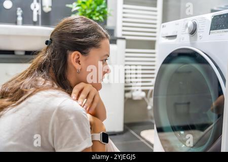 Jeune femme attendant que la machine à laver finisse, regardant la porte de la machine à laver Banque D'Images