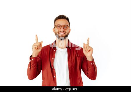 Bel homme arabe ou caucasien positif avec des lunettes, vêtu d'une chemise décontractée rouge, debout sur fond blanc isolé, montre les doigts vers le haut, à un espace de copie et de la publicité, sourit heureux Banque D'Images