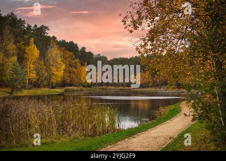 Scène automnale sereine avec un sentier paisible le long d'un lac tranquille entouré d'un feuillage d'automne vibrant, sous un ciel coloré de coucher de soleil Banque D'Images