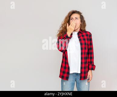 Femme bouclée en chemise rouge bâille sur fond gris Uni en studio. Banque D'Images