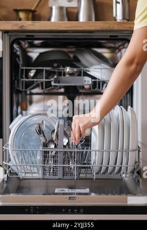 Vue avant de la main d'une femme enlevant les couverts propres d'un lave-vaisselle chargé. Fond de cuisine moderne Banque D'Images