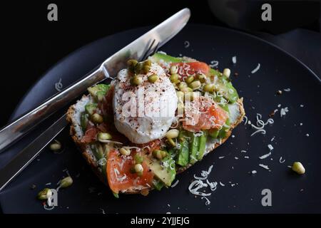 Oeufs pochés sur le dessus de l'avocat écrasé sur pain grillé au levain avec saumon fumé et germes de haricots mungo. Petit déjeuner sain avec assiette sur la table. Vue Angel. Banque D'Images