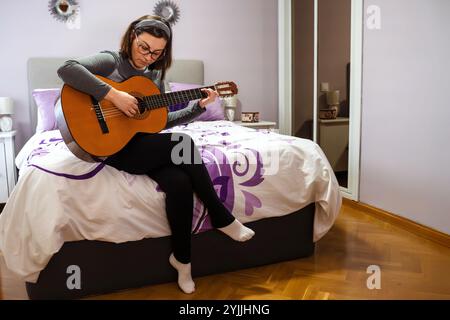 Femme assise sur un lit confortable dans une chambre violette, jouant de la guitare classique avec compétence et passion, entièrement immergée dans la musique Banque D'Images