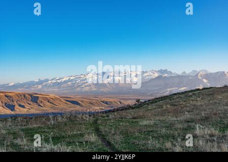 Une vue colorée à multiples facettes d'une colline envahie d'herbe jaune et d'un chemin, de collines sombres lointaines et de hautes montagnes enneigées sur un fond Banque D'Images