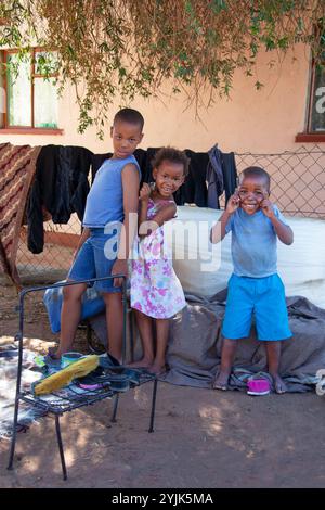 Trois enfants africains jouant dans la cour de la maison du village. Pauvreté, jouets fabriqués à partir de vieux meubles et objets. garçons et filles africains, Banque D'Images