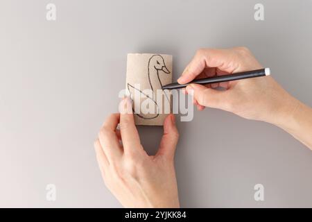 Vue de haut en bas des mains dessinant le cygne sur le rouleau de papier toilette vide, étape dans le processus créatif, figurine d'oiseau, concept de recyclage, bricolage pour les enfants, activité simple Banque D'Images