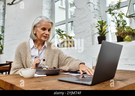 Une femme gracieuse aux cheveux gris est assise à une table, profitant de son repas tout en utilisant son ordinateur portable. Banque D'Images